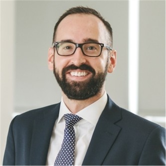 Callum Ross in glasses and a dark suit and tie, in front of a pale background